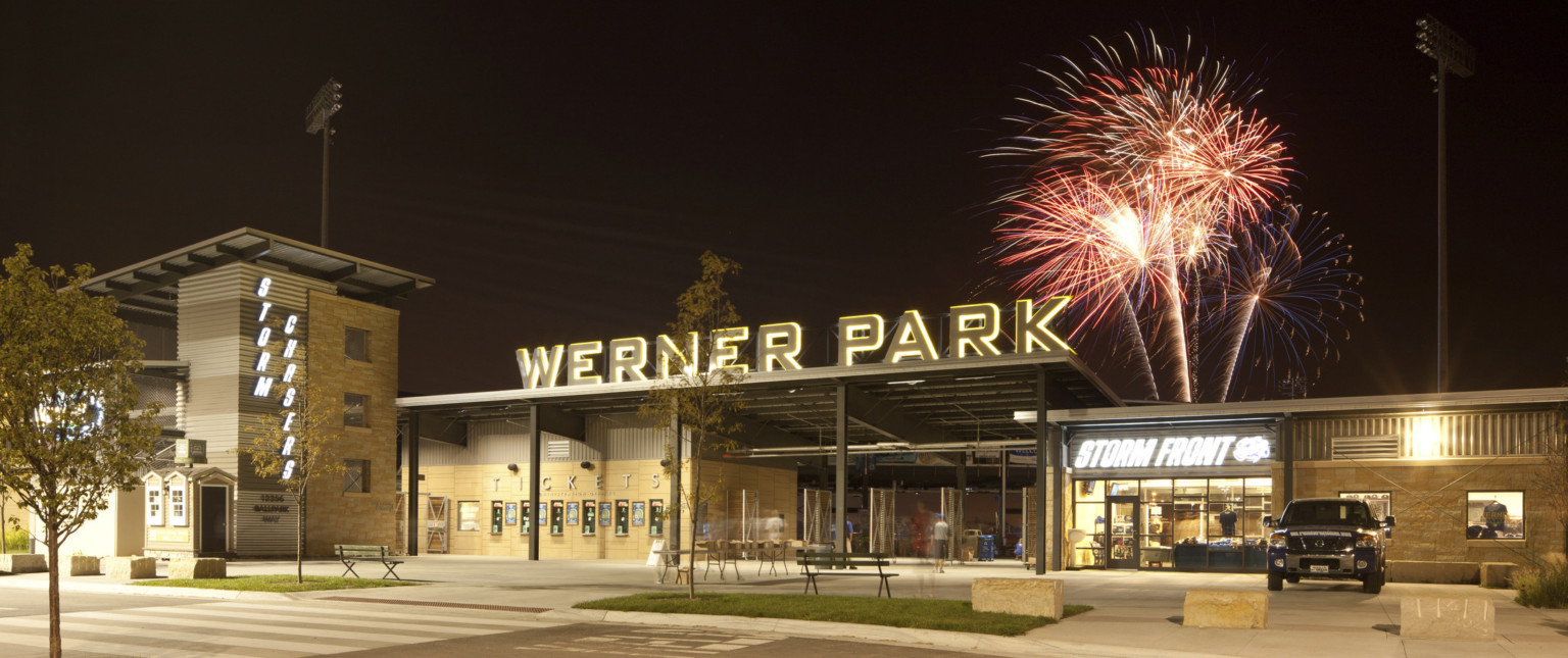 Stadium entrance with Werner Park sign illuminated at night. Right, Storm Front team store lit from within, fireworks above