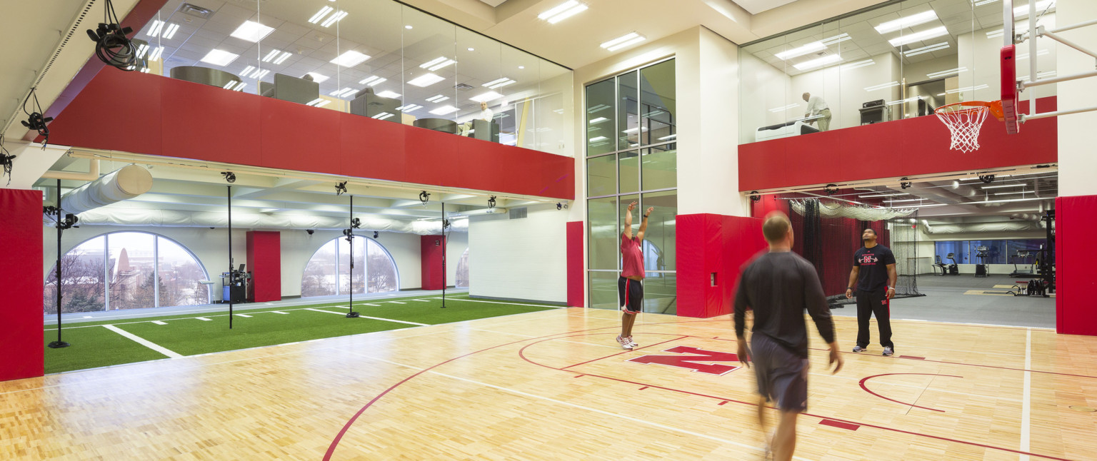 Double height basketball half court with windows on 2nd floor. Left, turf floor in front of arched windows with campus view