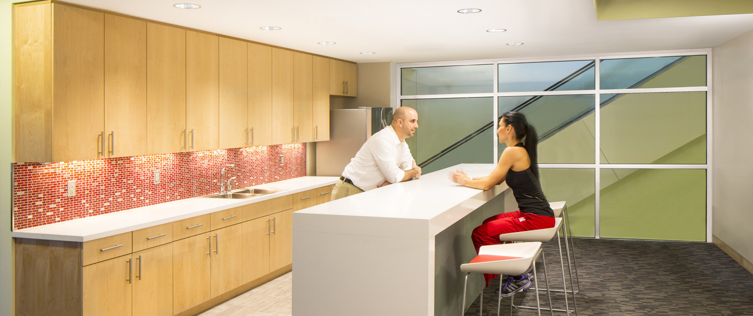 Kitchenette with red and white tiled backsplash between wood cabinets in white room. At center, island, with window, right