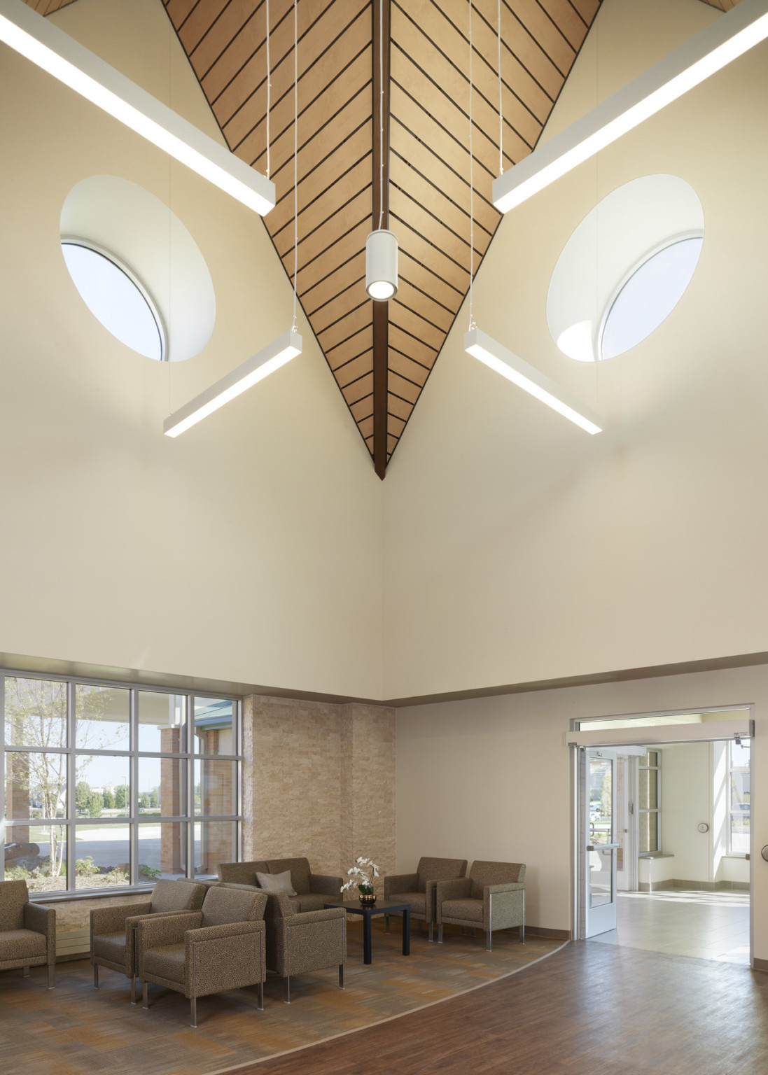 Corner view of beige room, with point of wood ceiling design descending corner between 2 oculus windows. Seating near windows