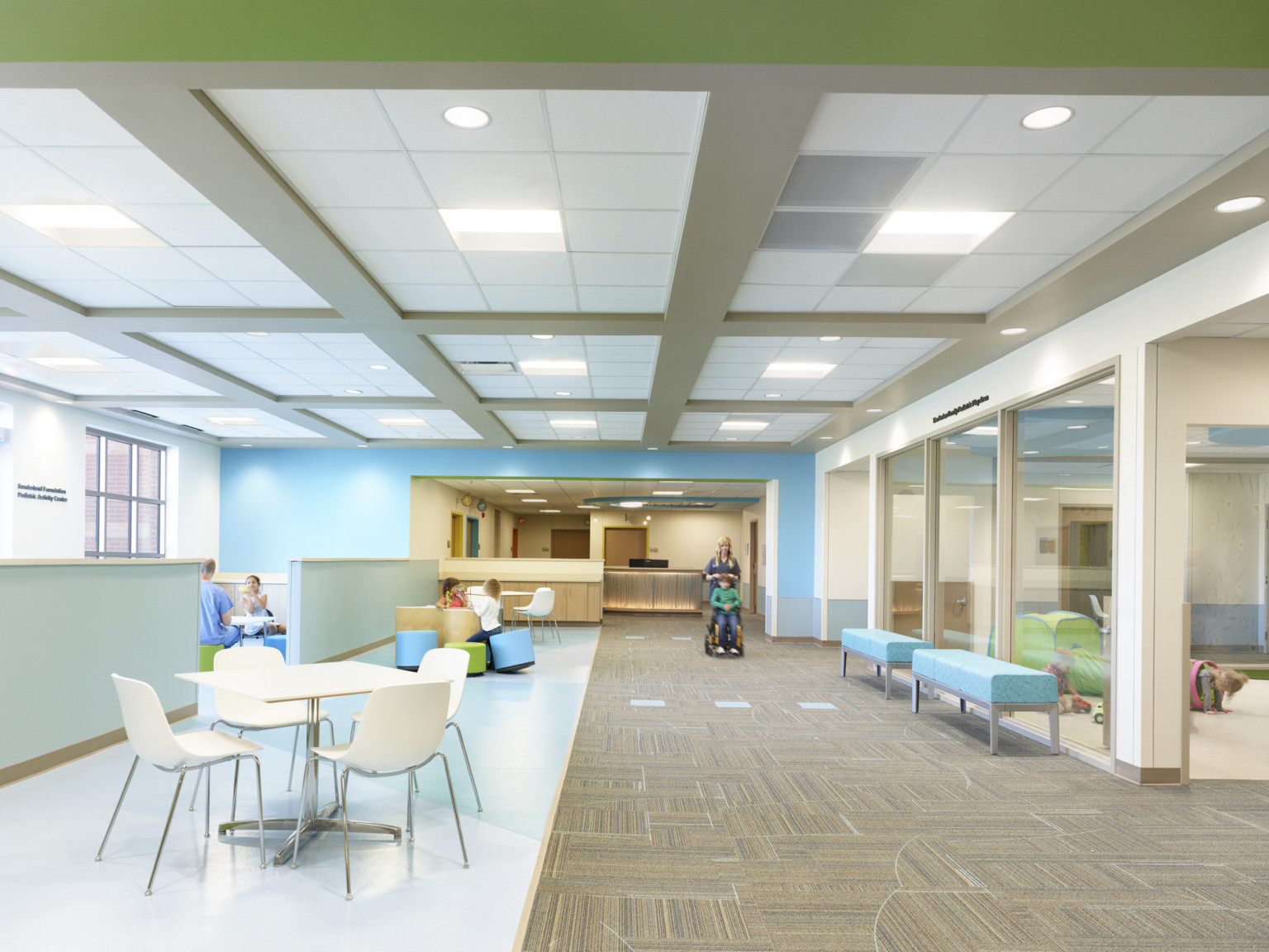 Right, carpeted hall with glass walled room lined with benches. Left is flexible seating in front of pastel blue half wall