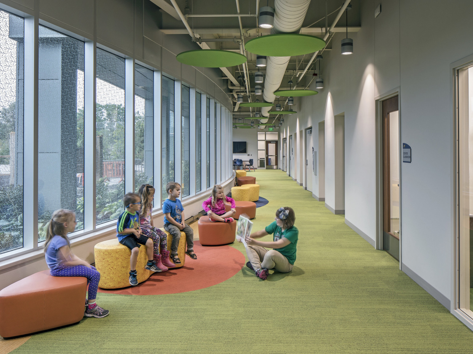 Hallway with large windows to the left lined with colorful stools. Green circles hang from above.