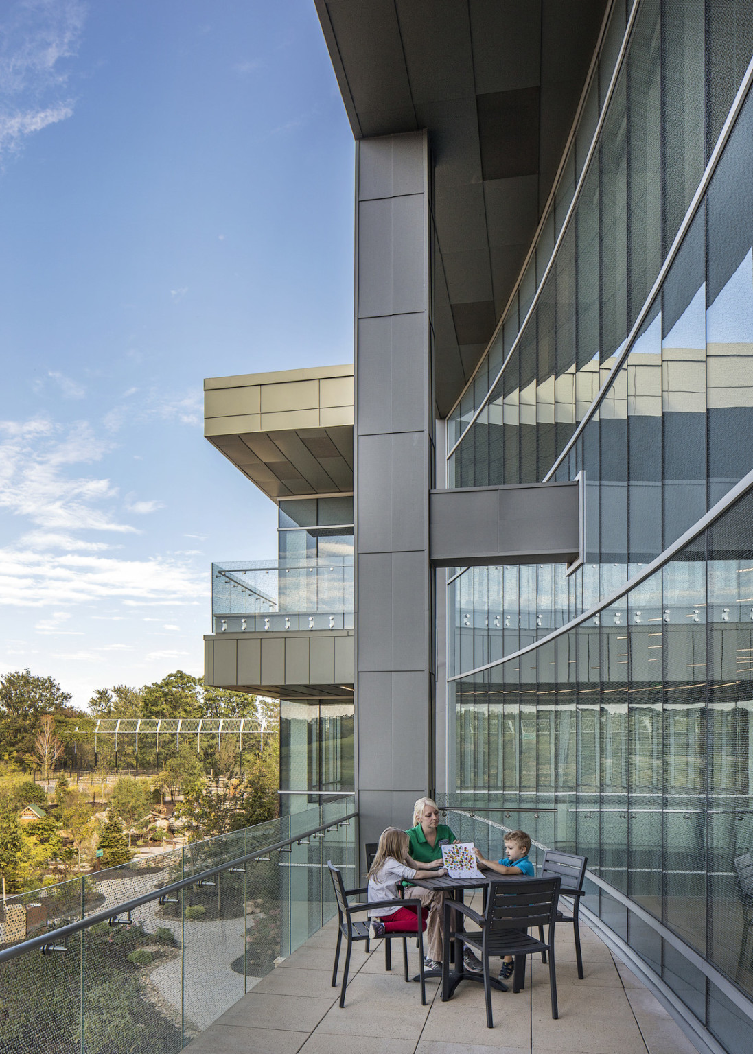 Curved wall of windows to right side of a glass walled balcony with patio furniture. A grey pillar at back connects to roof