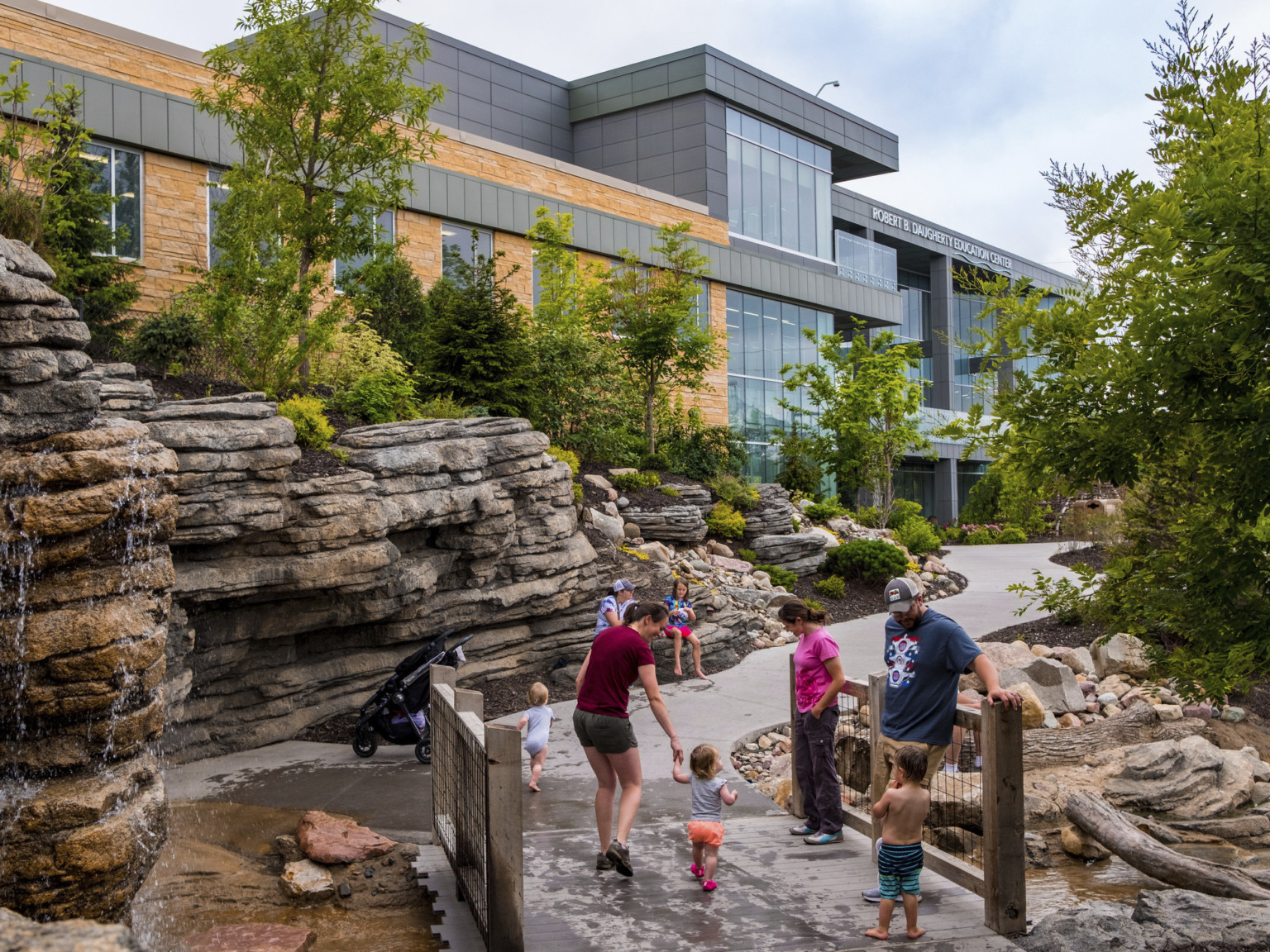 Small bridge over creek with waterfall flowing down rocks on the left along pathway leading to stone and glass building