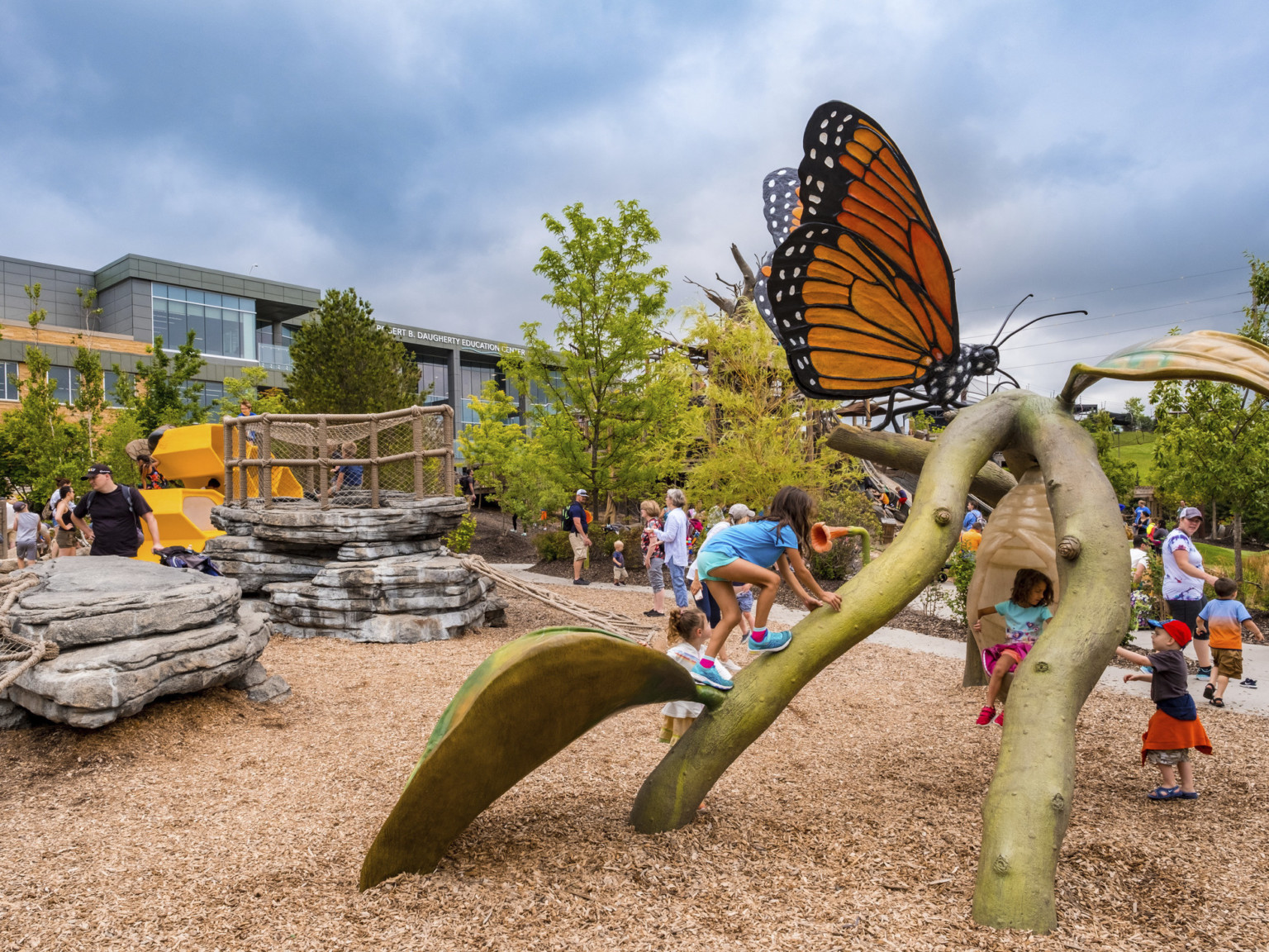 Playground with climbable sculpture of branch with leaves and butterfly with cocoon hanging down. Other equipment behind