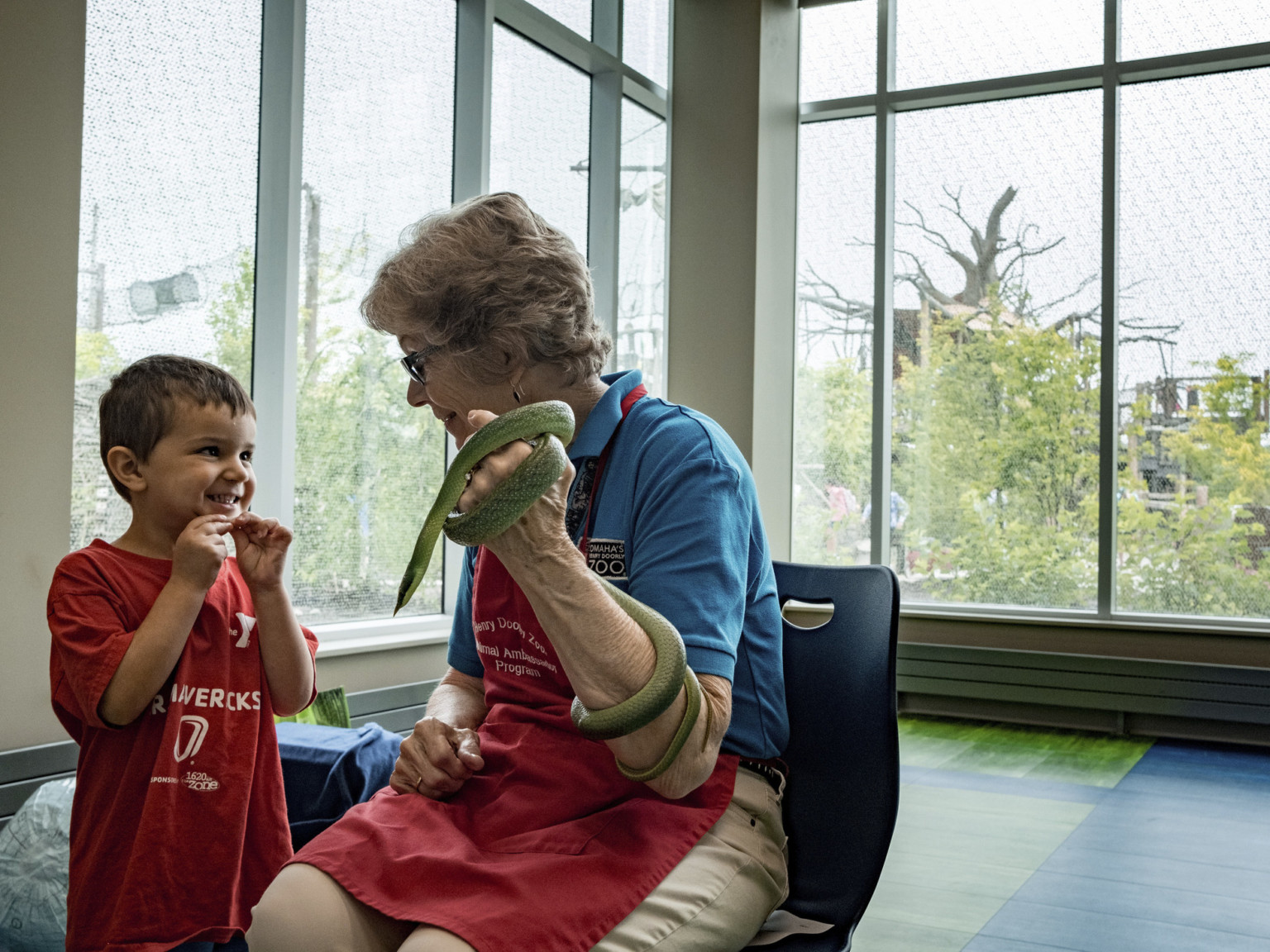 A woman holds a snake while speaking to a young boy in a room with green and blue flooring and large windows