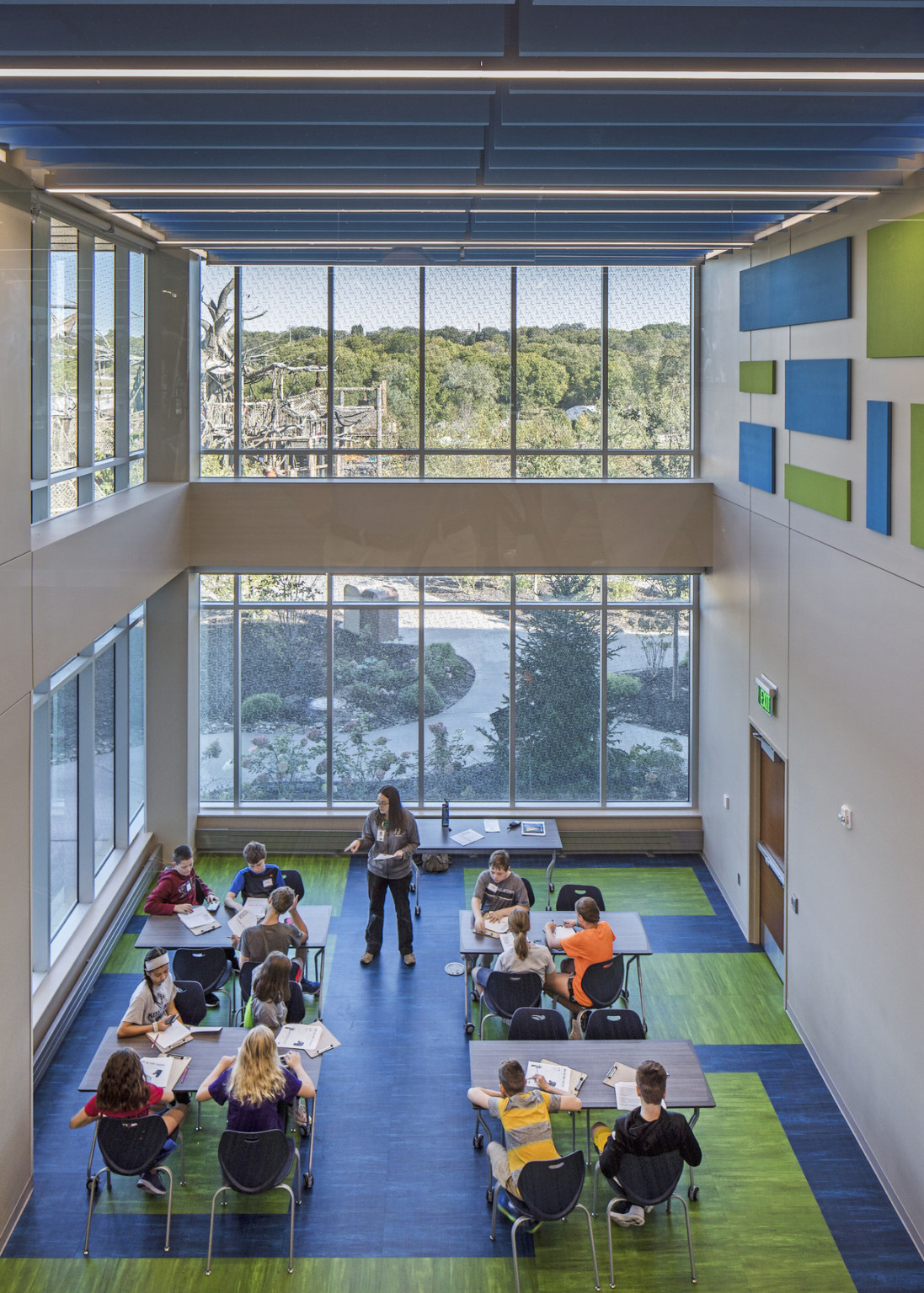 Double height classroom with window lined walls. Tables sit on green and blue carpet, these rectangles repeat on top of wall