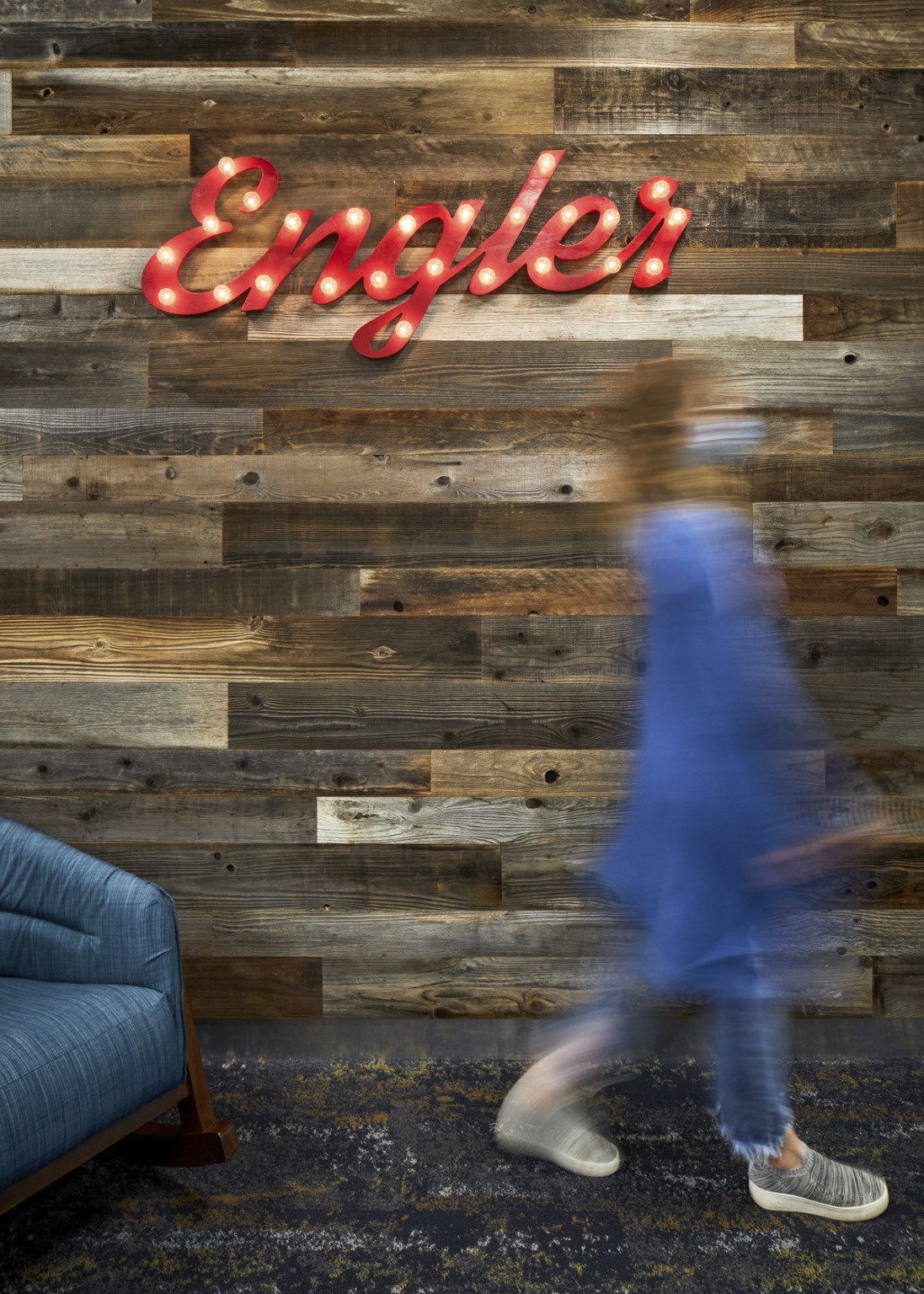 Wood panel wall with red script sign reading Engler, lit with small bulbs. Below, a blue armchair on carpet