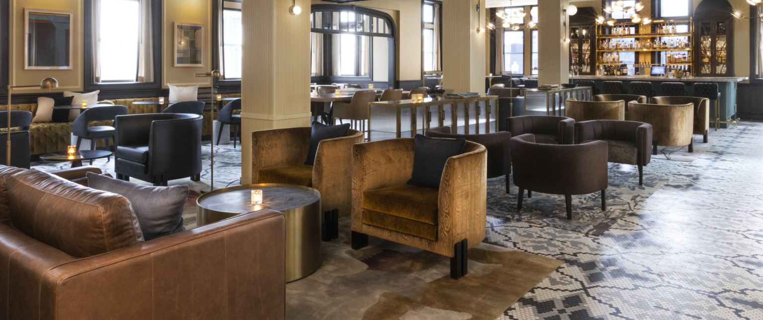 Seating area with arm chairs and round coffee tables and bar at back. Beige pillars below white ceiling with dark wood beams