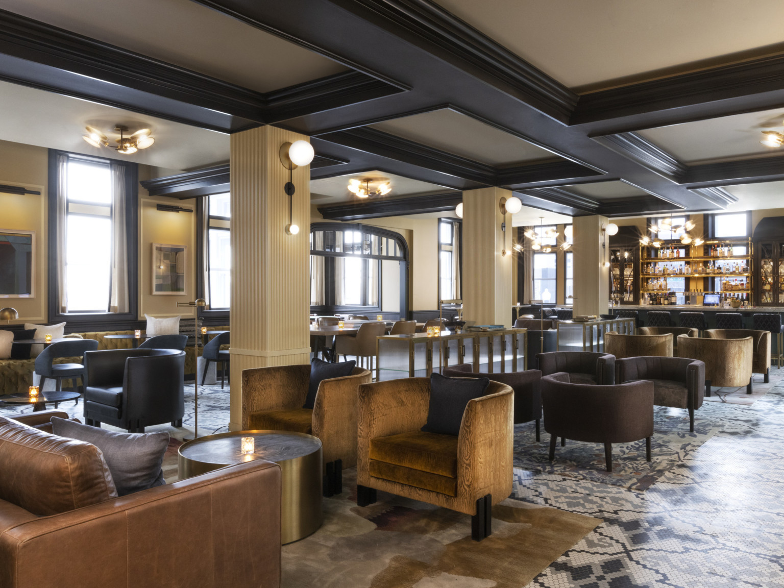 Seating area with arm chairs and round coffee tables and bar at back. Beige pillars below white ceiling with dark wood beams