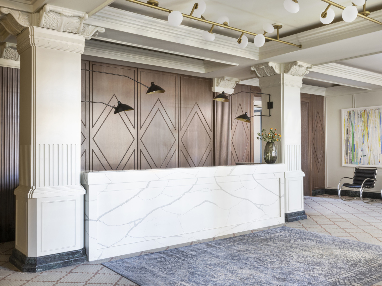 Lobby with white marble reception desk between Corinthian columns in front of geometric wood wall. Blue rug in front of desk