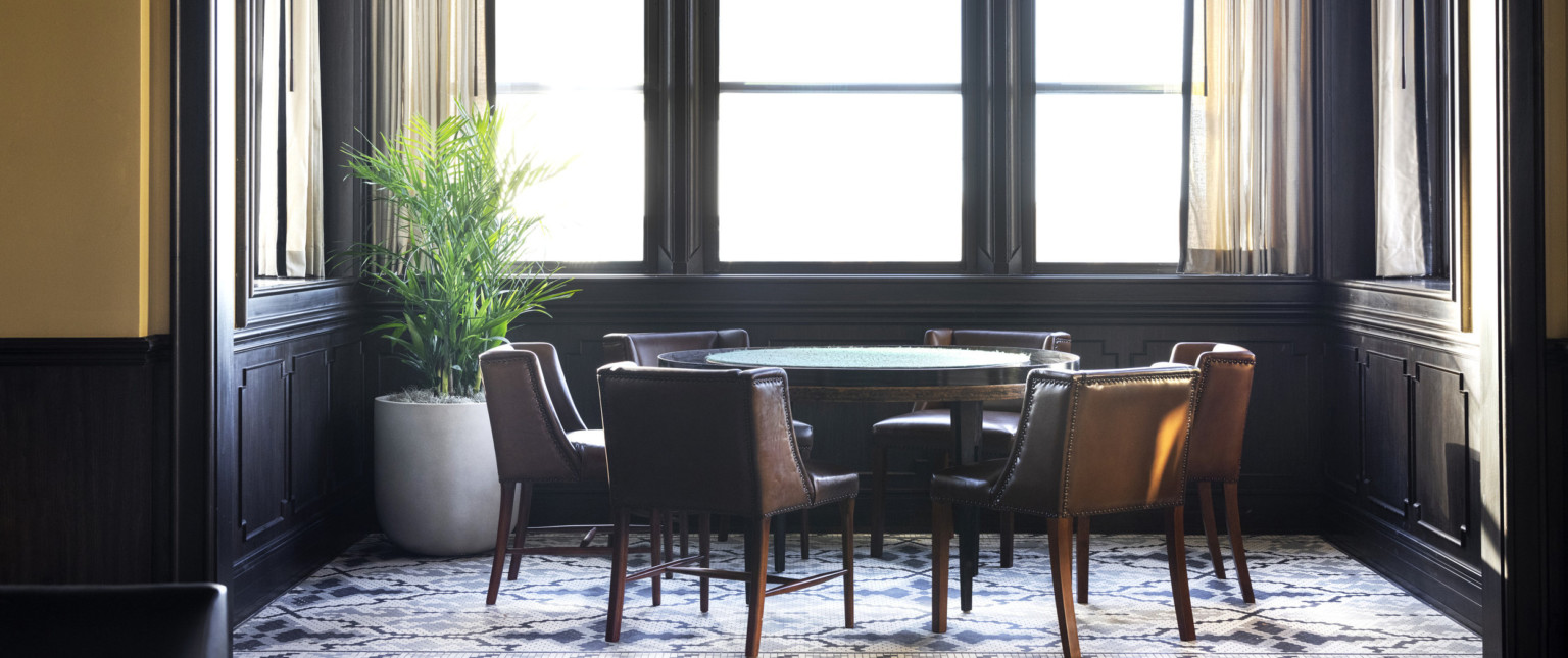 Poker table with 6 leather chairs in dark wood side room with large windows. Geometric flooring, and large potted plant left