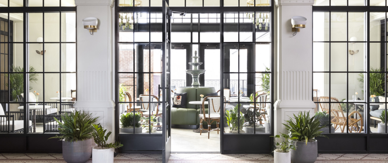 Floor to ceiling windows with door opened to seating area with fountain in round sofa. Room framed by white columns and plants