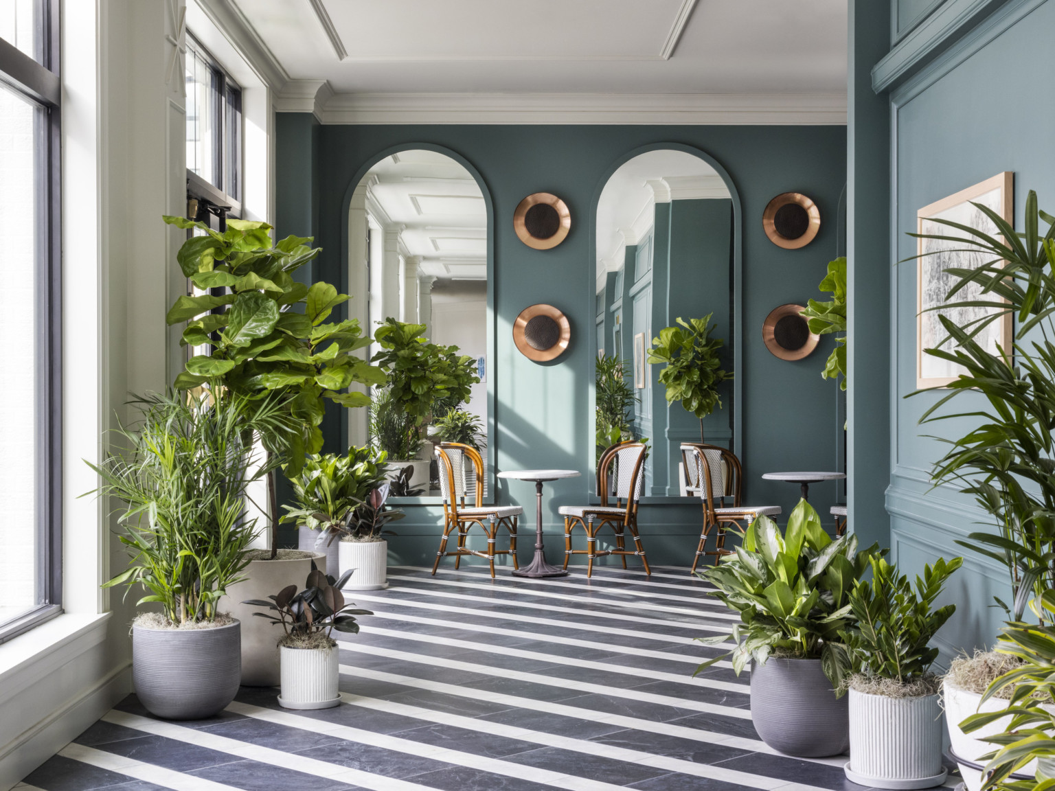 Black and white diagonal striped floor in green window lined hallway. Seating area and arched mirrors at end of hall