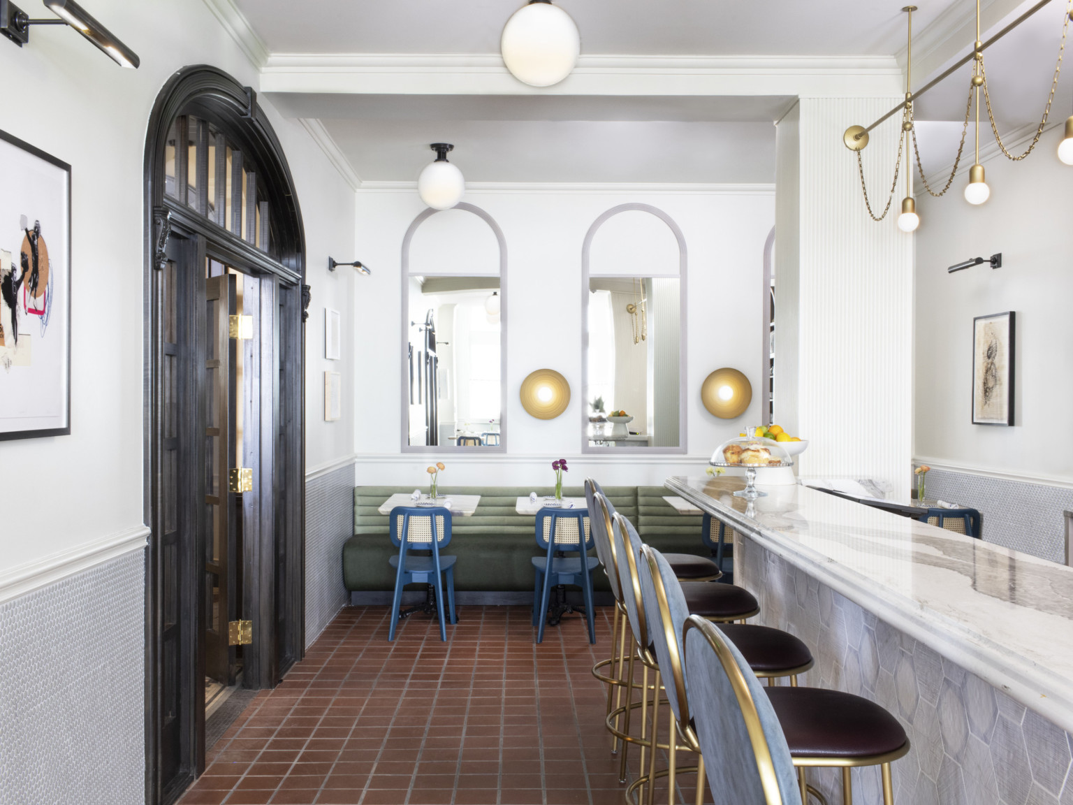 White room with dark wood arch doors left. Marble counter, right, with blue stools. Small tables behind with arched mirrors