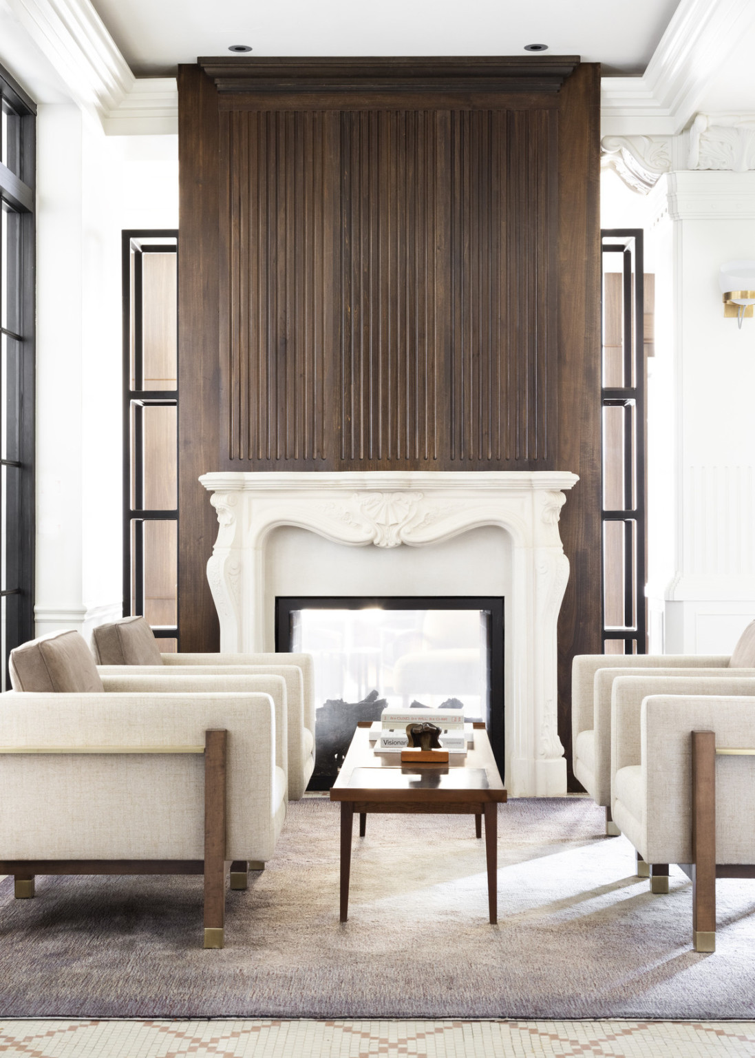 4 arm chairs around long wood coffee table on rug in front of carved white stone fireplace with wood overlaid chimney