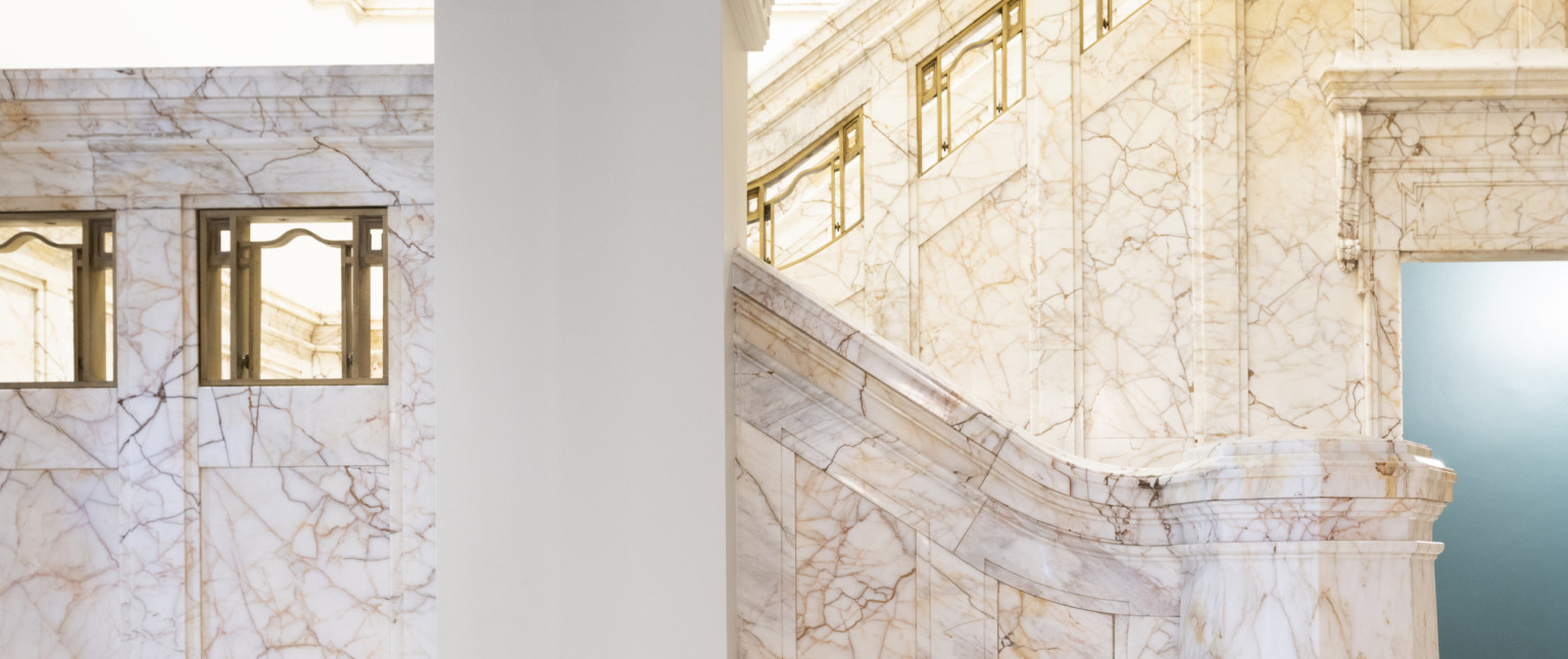 White column in a marble staircase with curving rail with sections of golden metal work inset