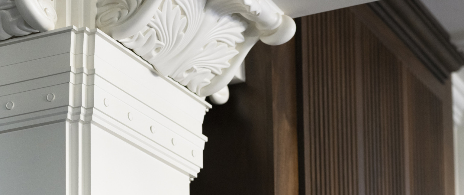 Close up of organic carving in capital of white stone Corinthian column in front of dark wood paneling