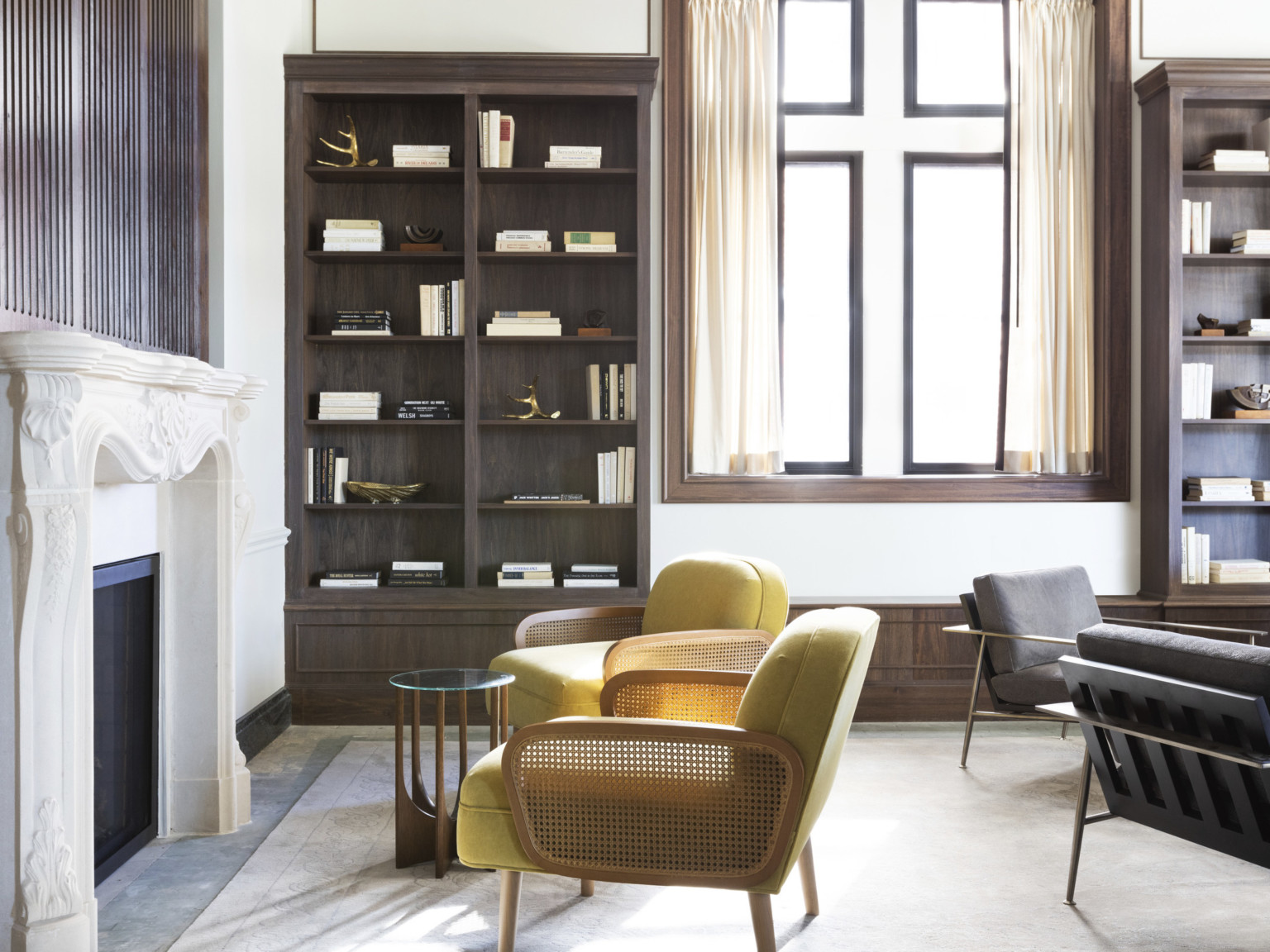 Seating area with arm chairs and table in front of white stone carved fireplace. Dark wood built in bookshelves beside window