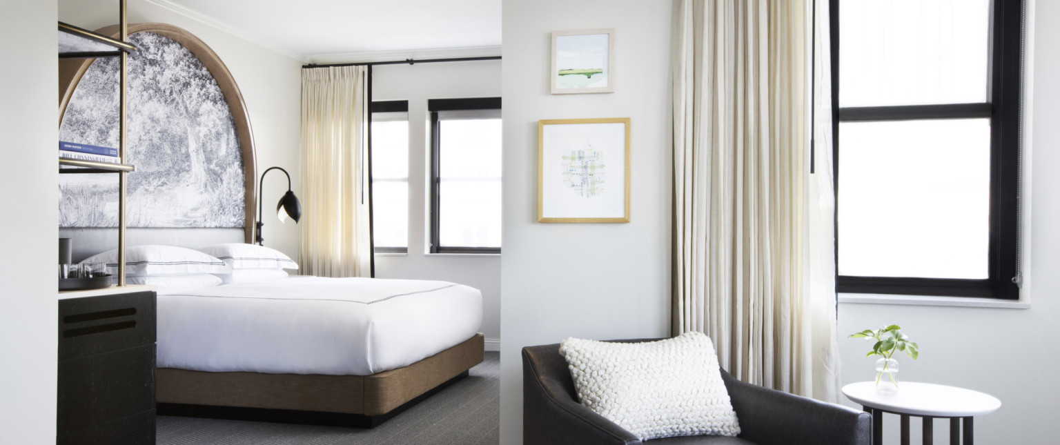 Black arm chair with white knitted pillow in sitting area in front of bedroom with white bed on wood base and large windows