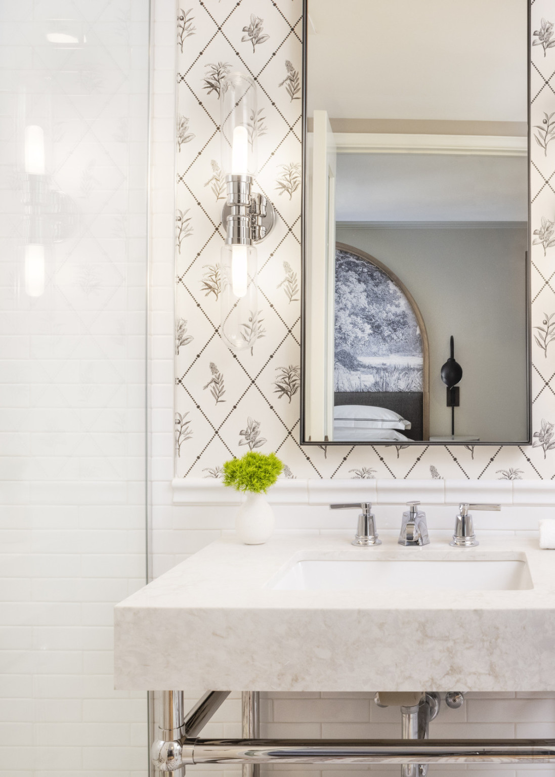 White marble sink on white tiled wall with black and white floral wall paper detail above. Mirror over sink reflects the bed