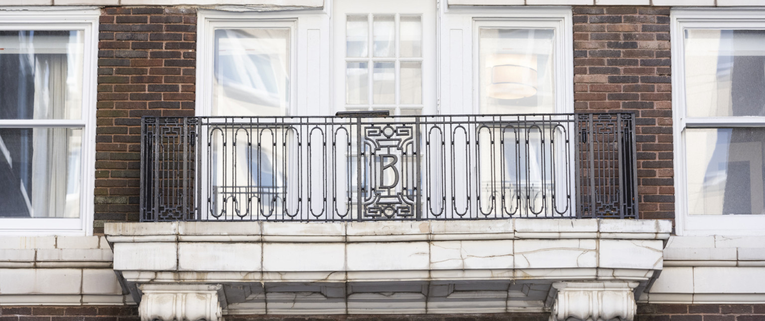 Balcony at window with white stone base and wrought iron bannister, the letter B framed at center