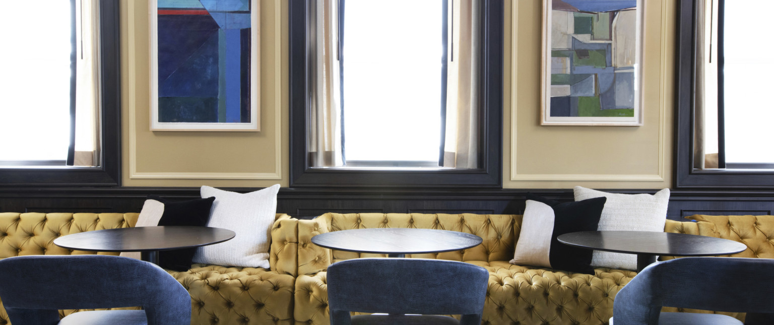 Yellow tufted booth along dark wood base board on beige wall. Black and white pillows on booth with round tables and chairs