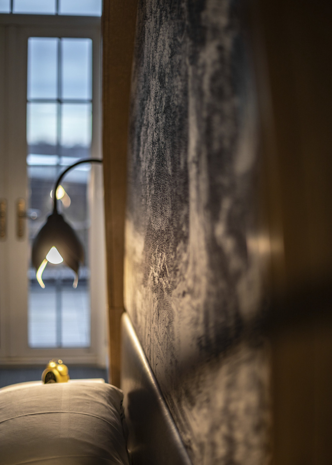 Close up of black and white muraled headboard in evening, illuminated by organic, tulip shaped black bedside lamp and window