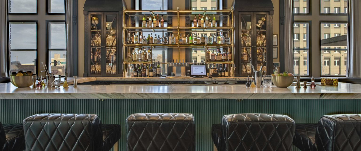 Close up of bar with white marble counter and green ribbed side. Gold toned shelves above back of bar, quilted seats in front