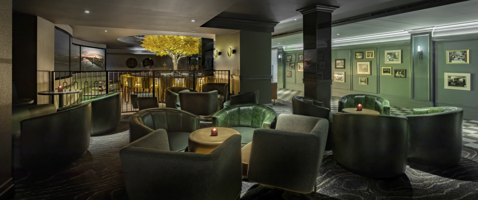 Offset seating area with arm chairs in green room, separated from bar by dark metal bannister. Behind, green hall with photos