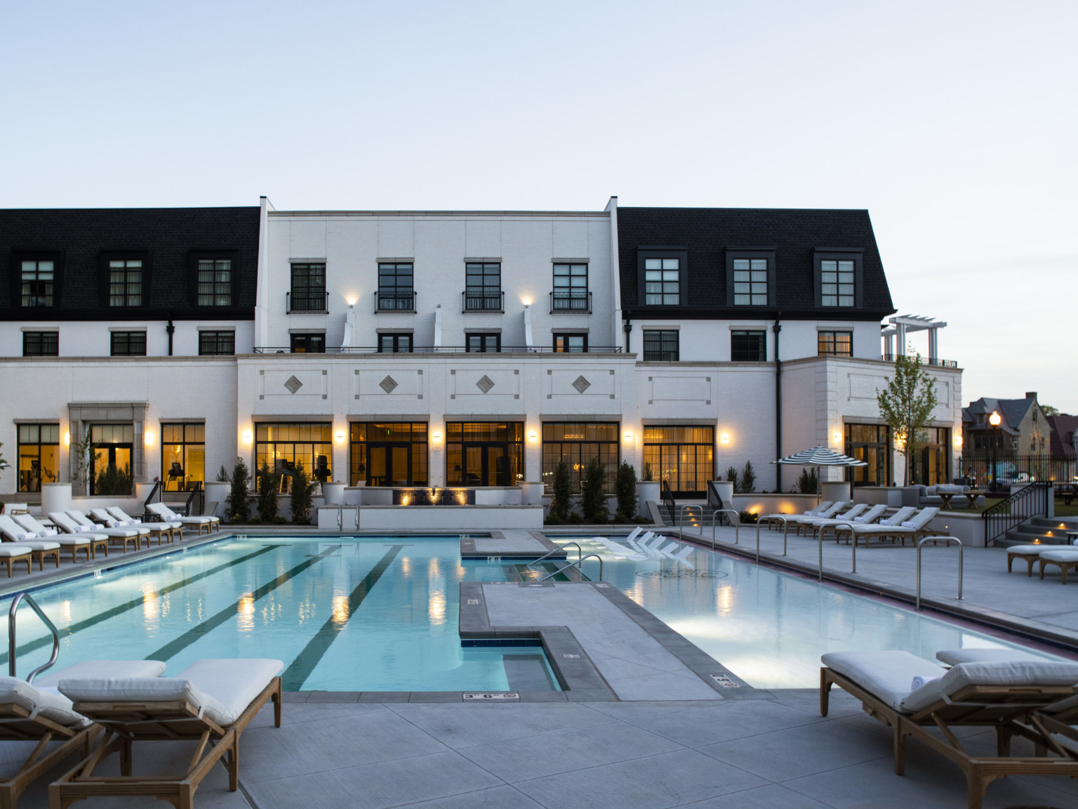 Outdoor pool surrounded by lounge chairs. White 3 story building behind, with floor to ceiling 1st floor windows and pillars