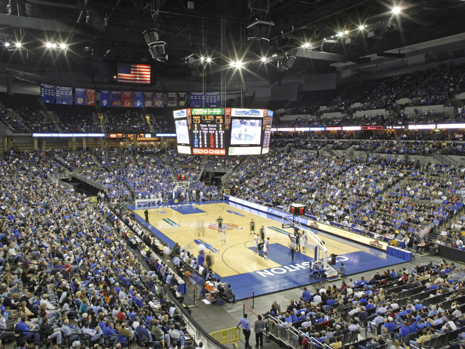 Basketball court with lighting and jumbotron hanging above. Bleachers surround the court and are filled with fans