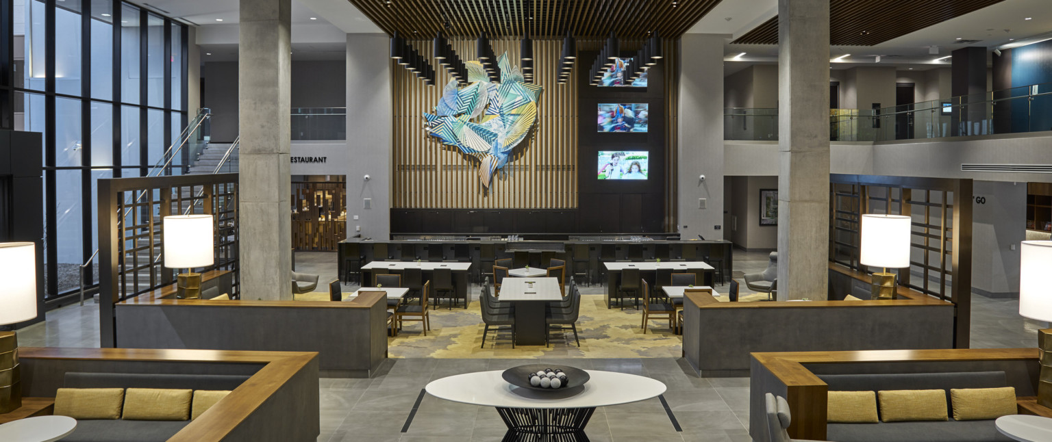 Mezzanine hallway in atrium with bar and tables at front of lower level. Stone pillars center and side 2nd floor walkways