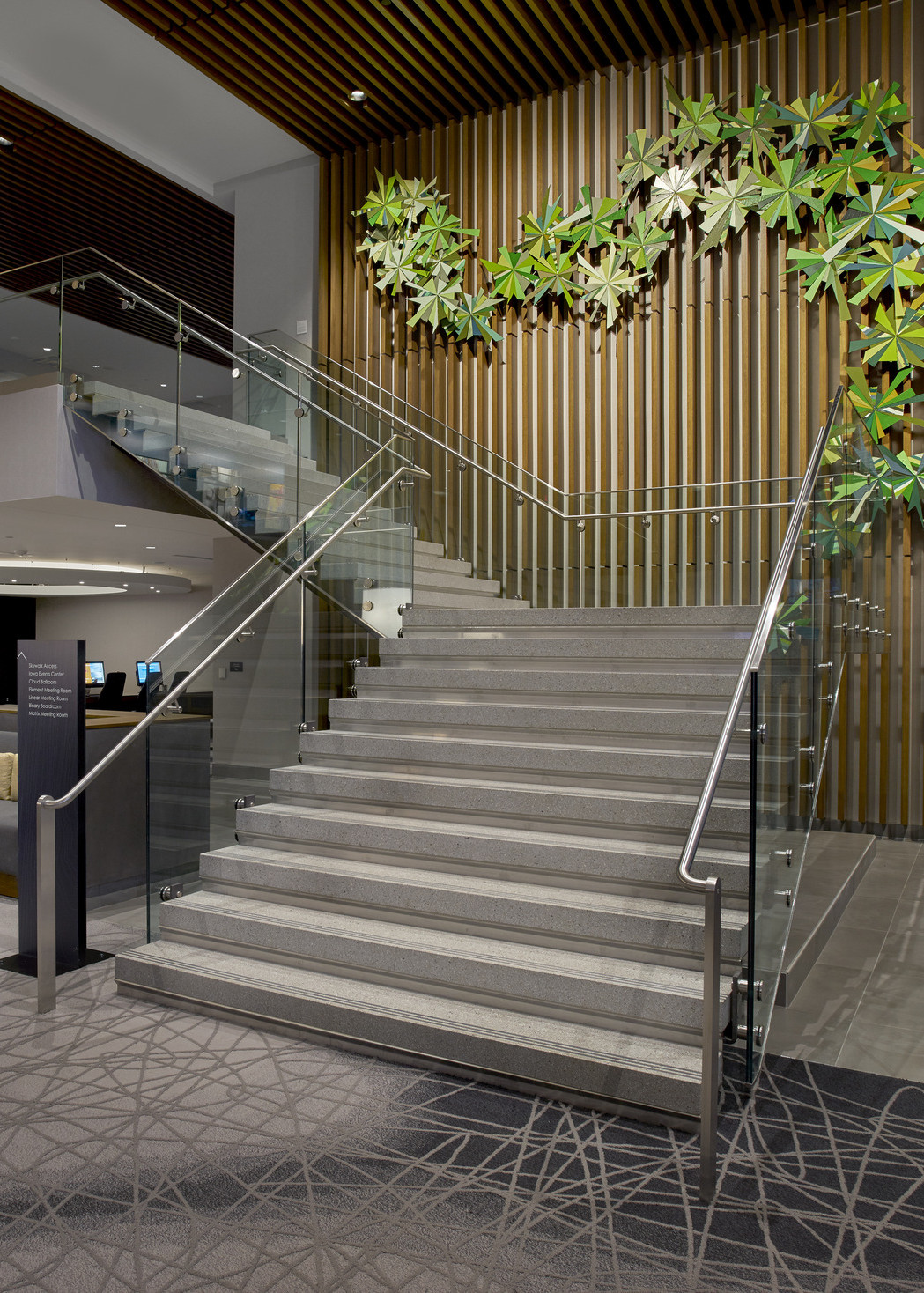 Grey stairs with glass railing, a ribbed brown wall to the side with organically shaped green sculpture curling up the wall