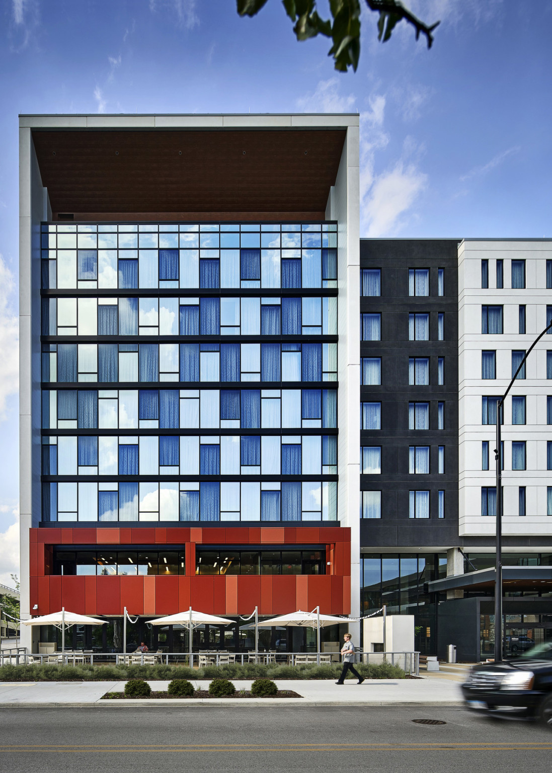 6 story window wall left above red portion of building behind an outdoor patio with dining space, tables, and umbrellas