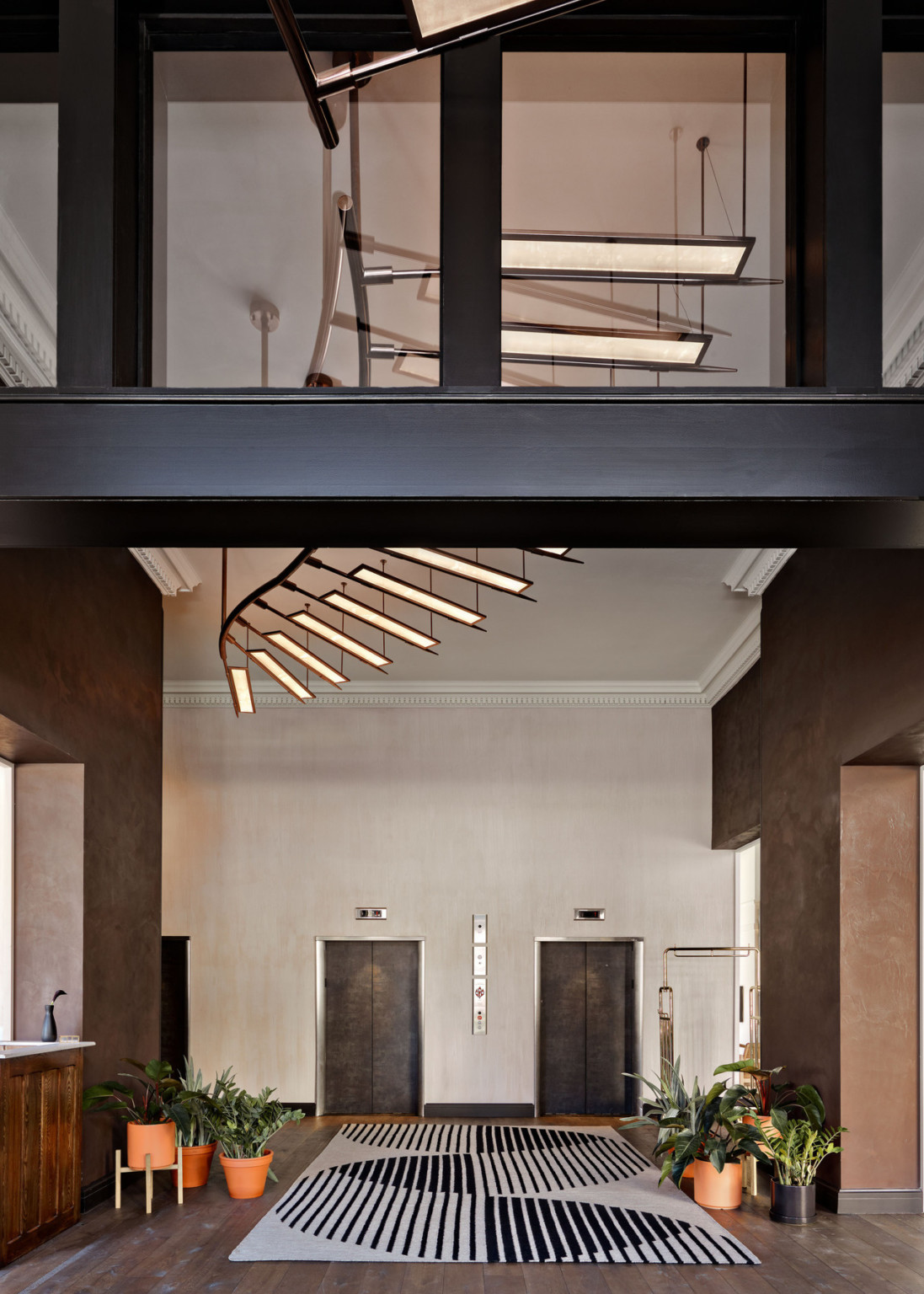 Spiraling paneled light fixture over double height elevator room with geometric grey and black carpet. Plants on each side