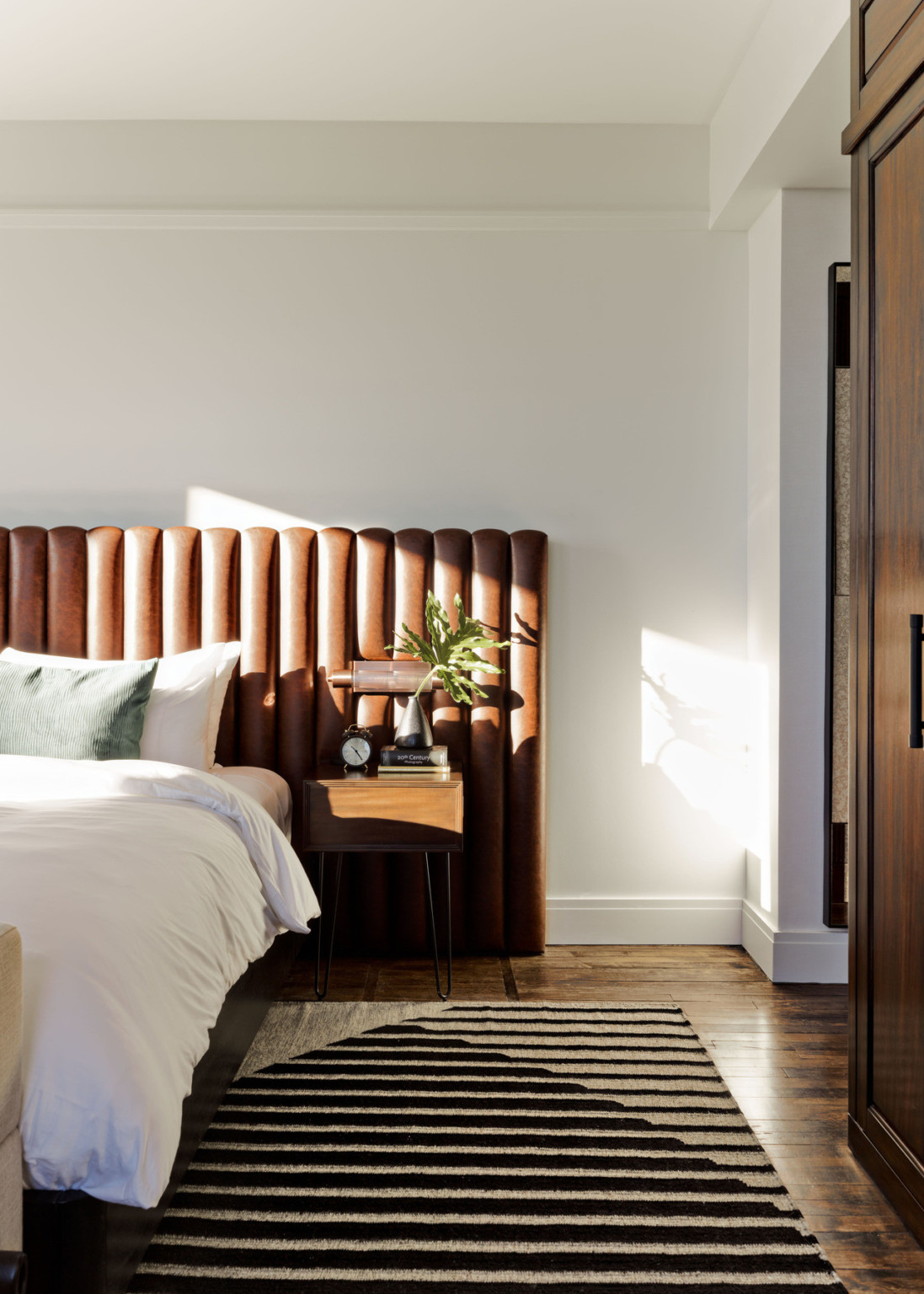 White bed with brown panel upholstered headboard in white room with wood side table over black and grey geometric striped