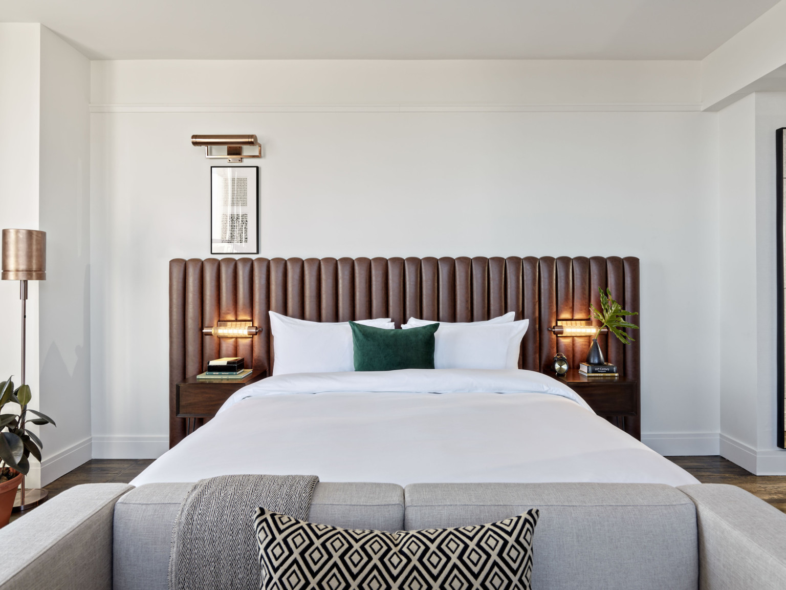 White bed with green throw pillow against a brown ribbed headboard. Side tables and lamps on each side, grey couch at base
