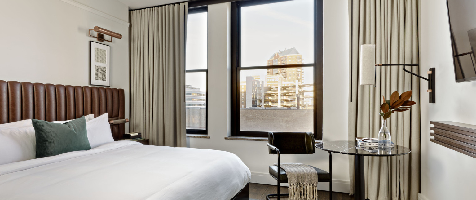 White room with bed with brown headboard, round black table and black cesca chair next to window with view of tall buildings.