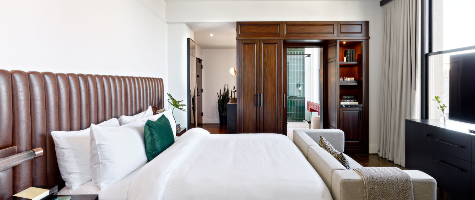 Bedroom with white bed with grey couch at the foot, brown wood built in cupboard and shelves framing pocket door to bathroom