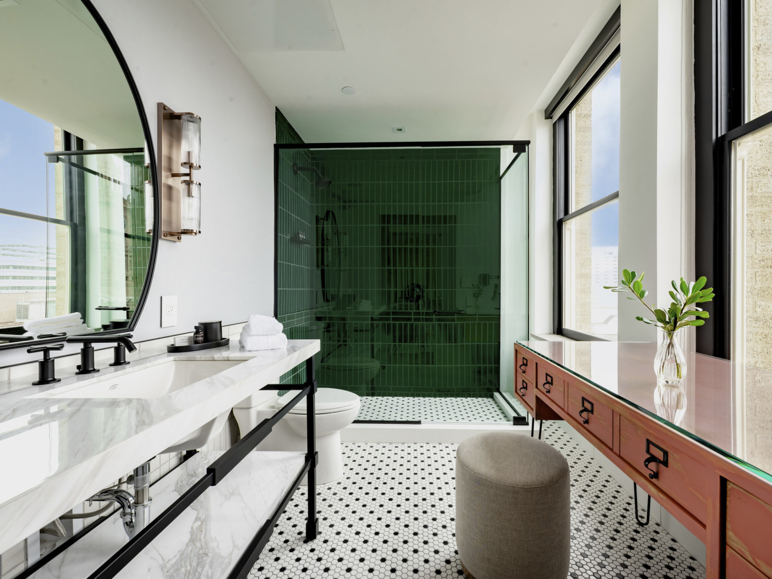 Bathroom with green tiled shower, toilet, and sink left. Right are 2 large windows in front of table and stool