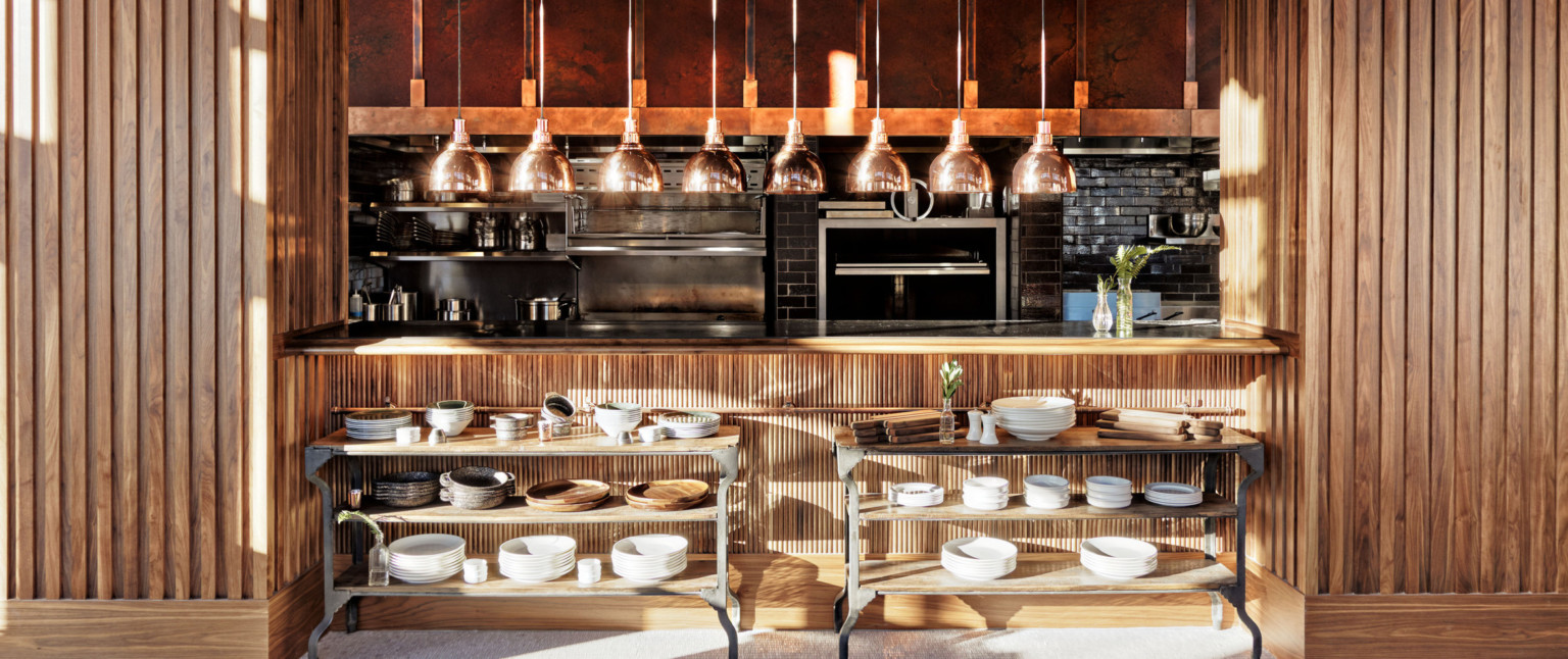 Wood walls and counter with cutout to exposed dark brick kitchen. Wood and metal shelves under counter, appliances behind