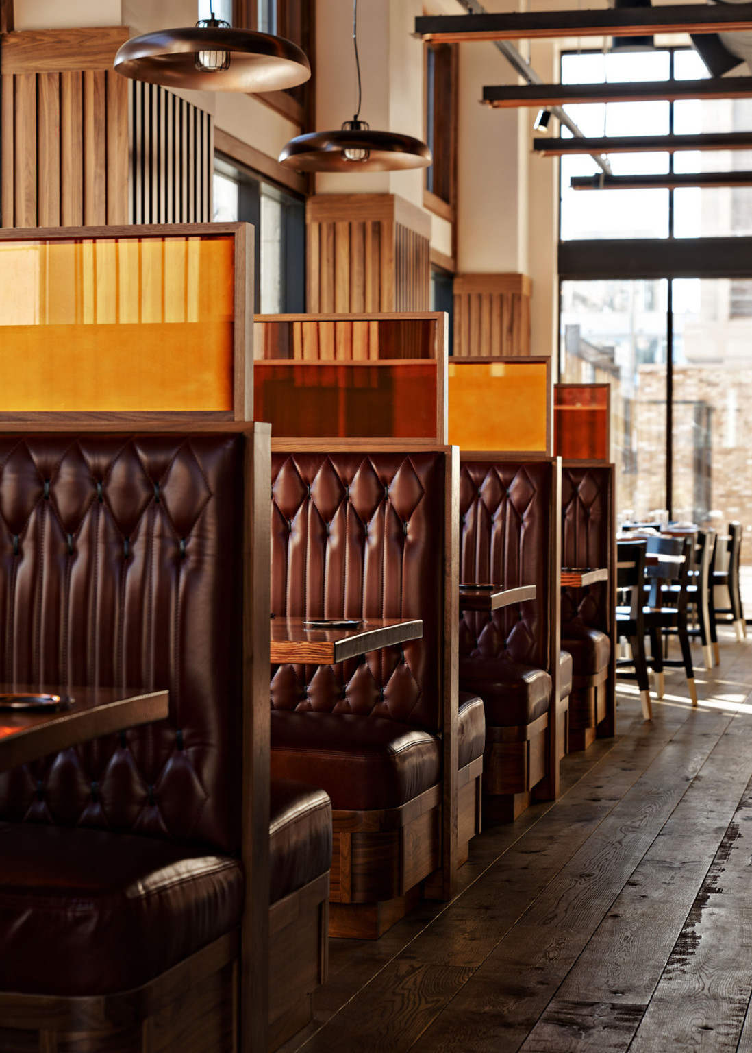 Dark quilted booths with dark wood accents along side of room with floor to ceiling windows and circular hanging lamps