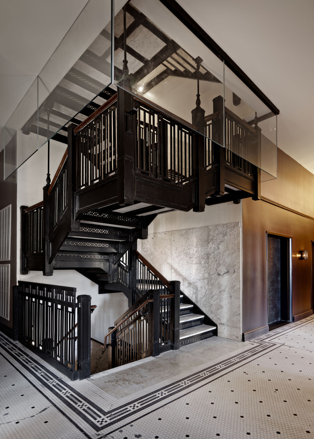 Black metal stairs with marble steps in white stairwell at the corner of a tiled hallway with wood walls.