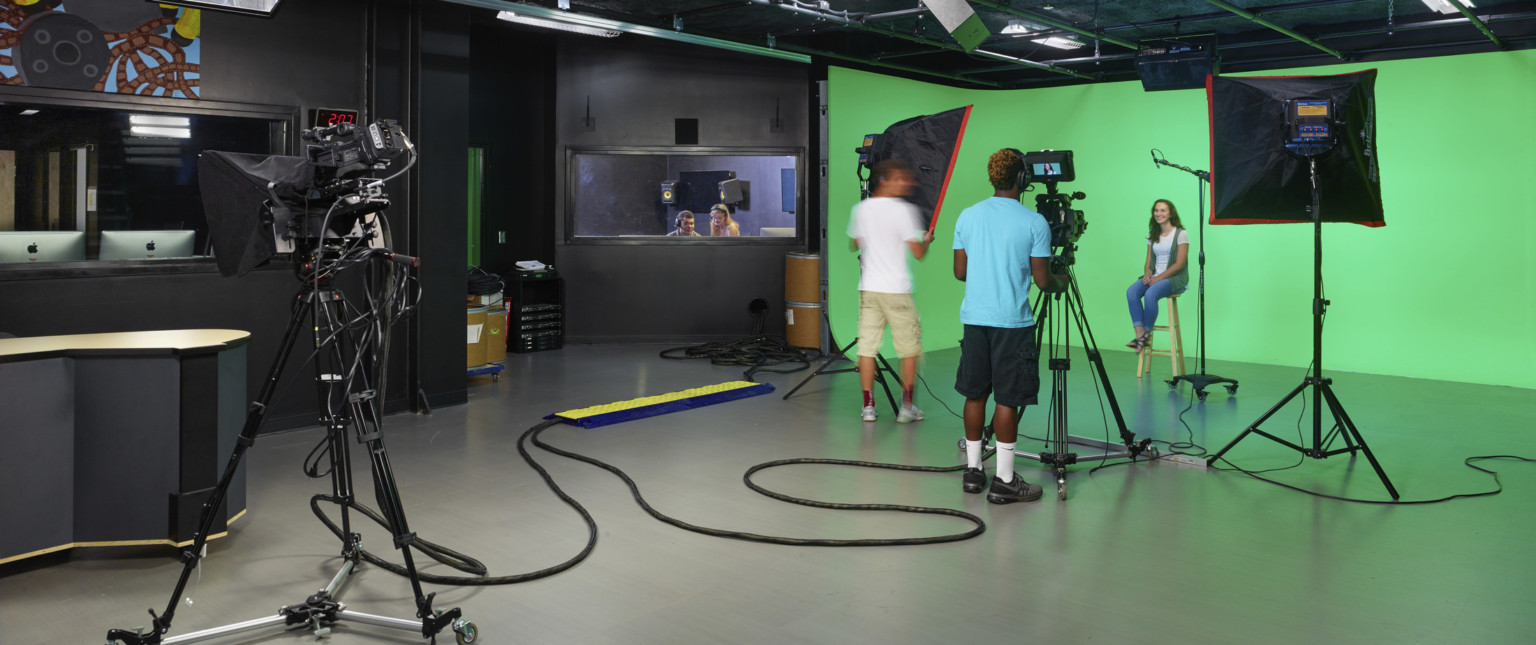 Students operate camera and lighting equipment in front of green screen in black room. Window, left, to editing room