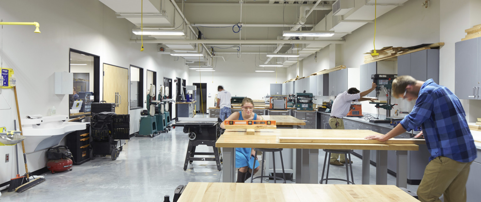 Students work in woodshop, a white room with grey cabinets to right. Power cords hang from ceiling with exposed ducts