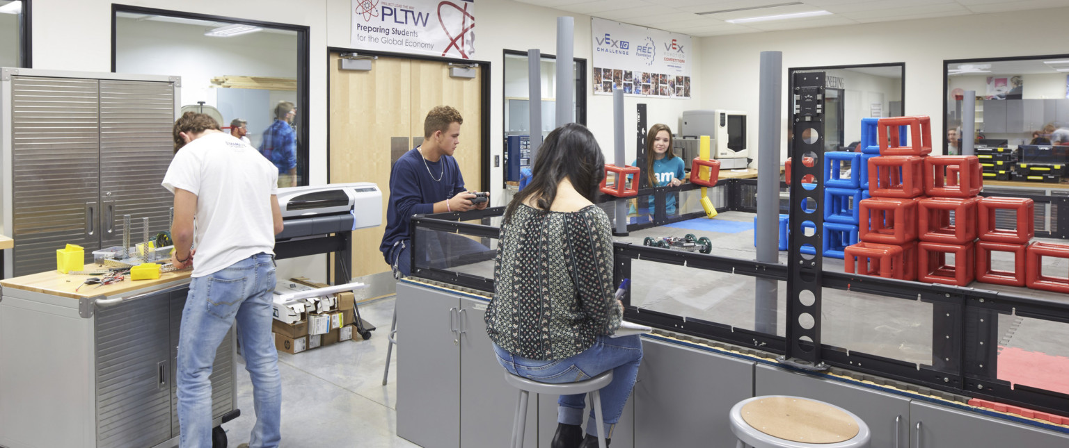 Students work in robotics lab. Large boxed in counter with stools has remote control car and blocks on top.