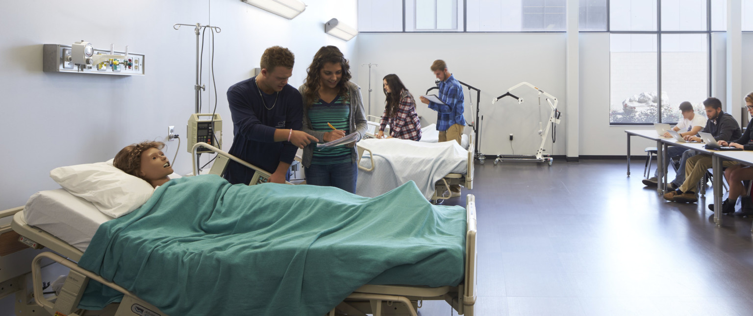 White room with high ceilings and windows on back wall. Hospital beds to left with CPR dummies. Tables face them at right