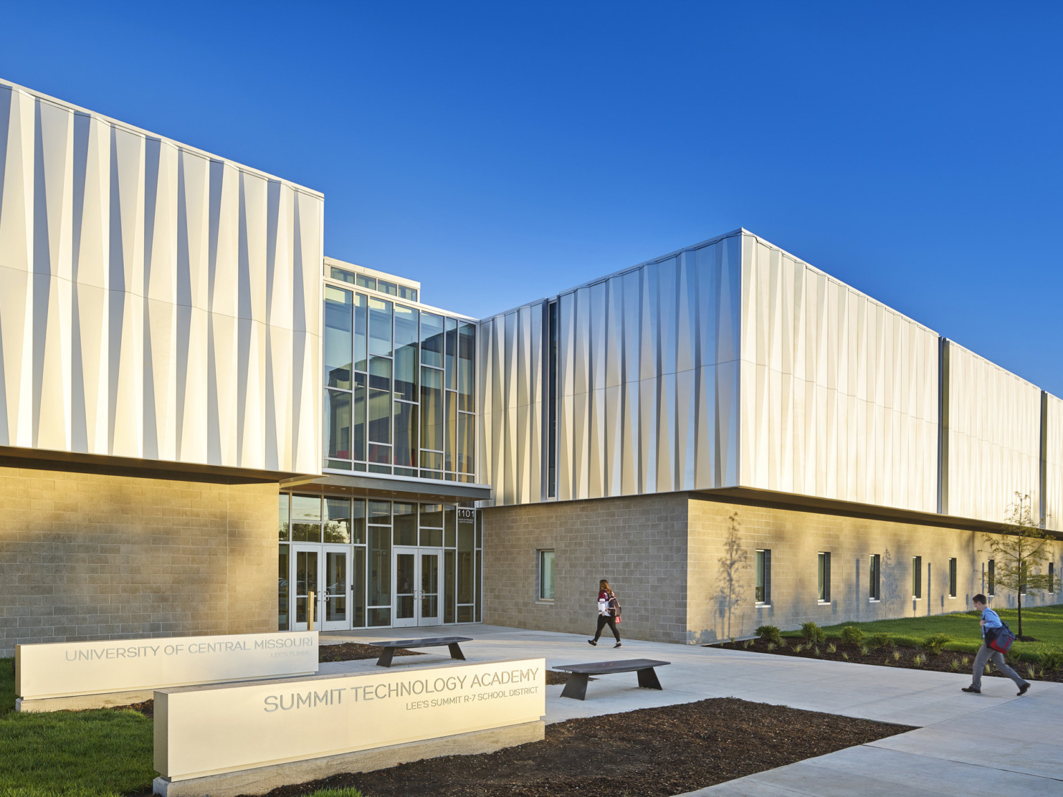 Exterior view of 2 story building with stone lower building and textural metallic upper. Double height windows at center