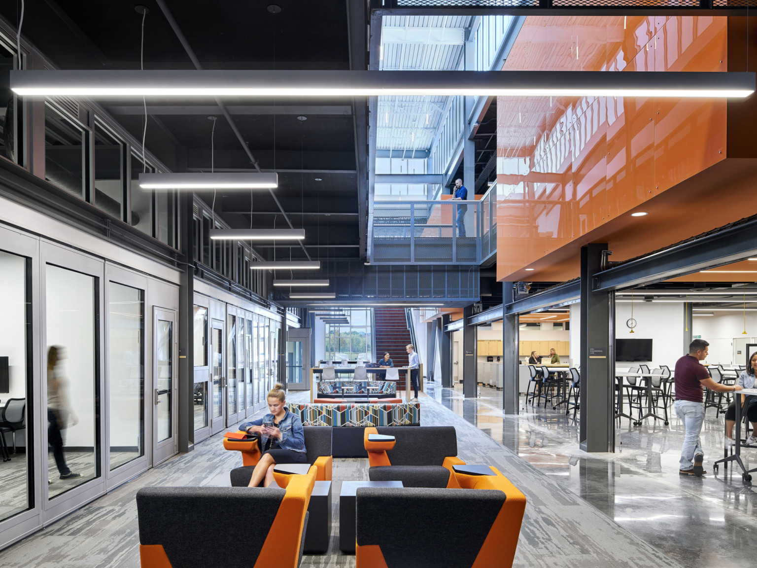 Atrium seating area divided by a hallway. Windows look into learning spaces, left. A 2nd floor walkway extends above left